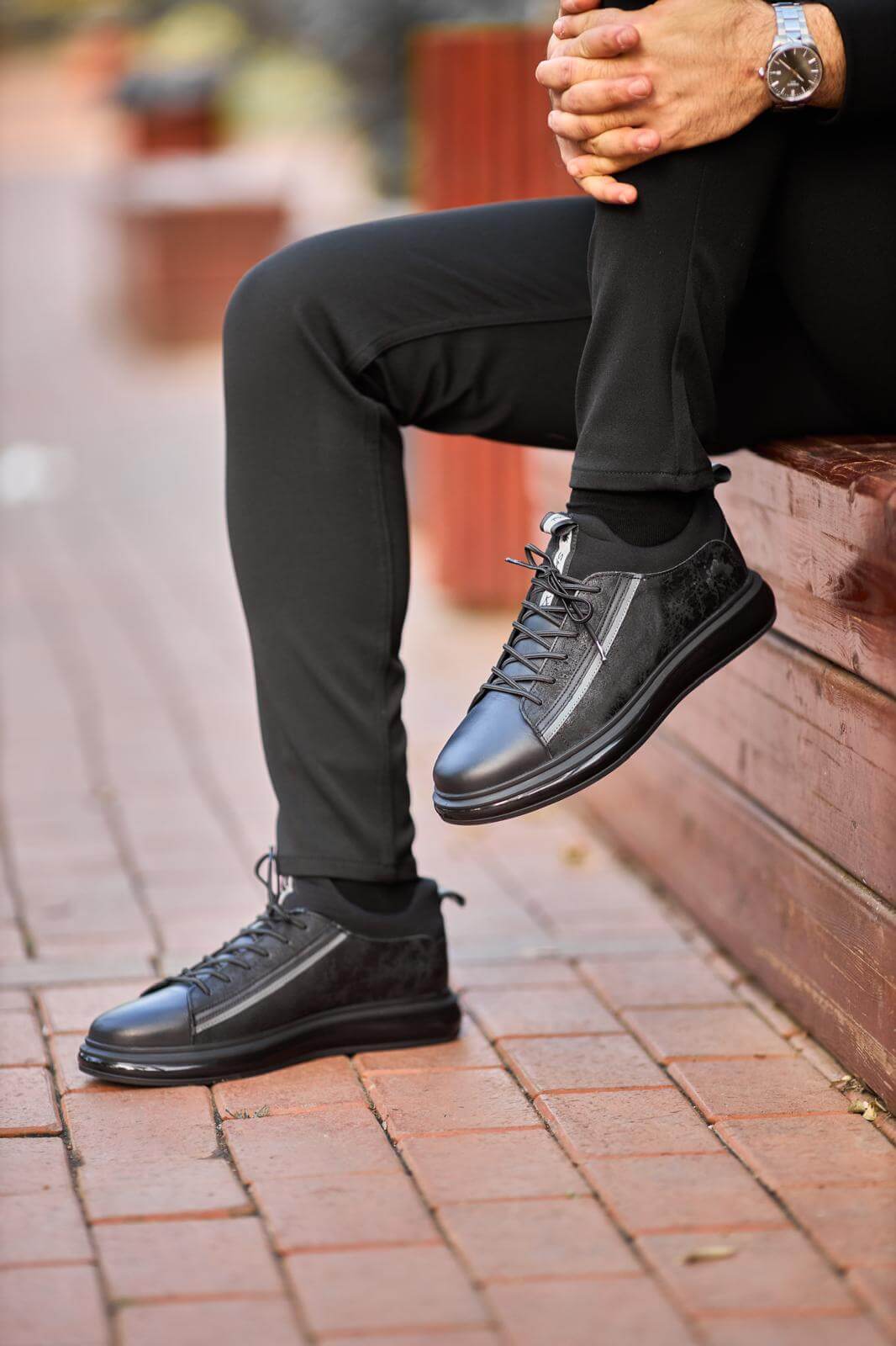 Black genuine leather sneakers with suede panels, worn with black trousers on outdoor wooden bench.