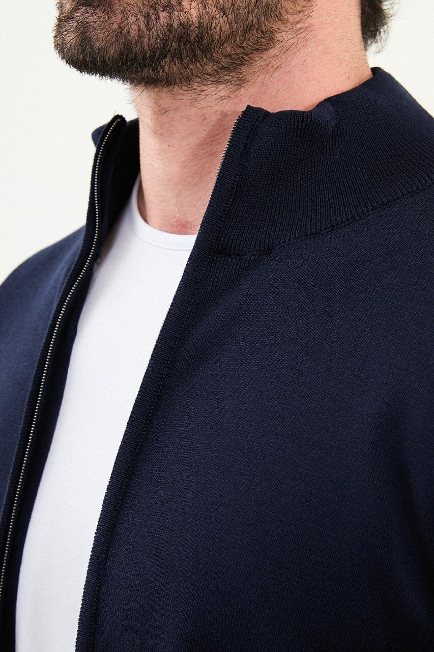 A man wearing a slim fit navy blue zip cardigan with a stand-up collar, styled with a white T-shirt and dark jeans.