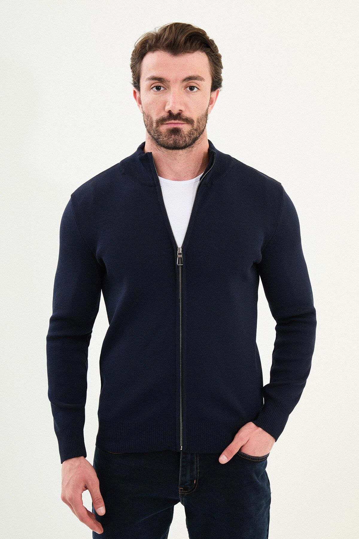 A man wearing a slim fit navy blue zip cardigan with a stand-up collar, styled with a white T-shirt and dark jeans.