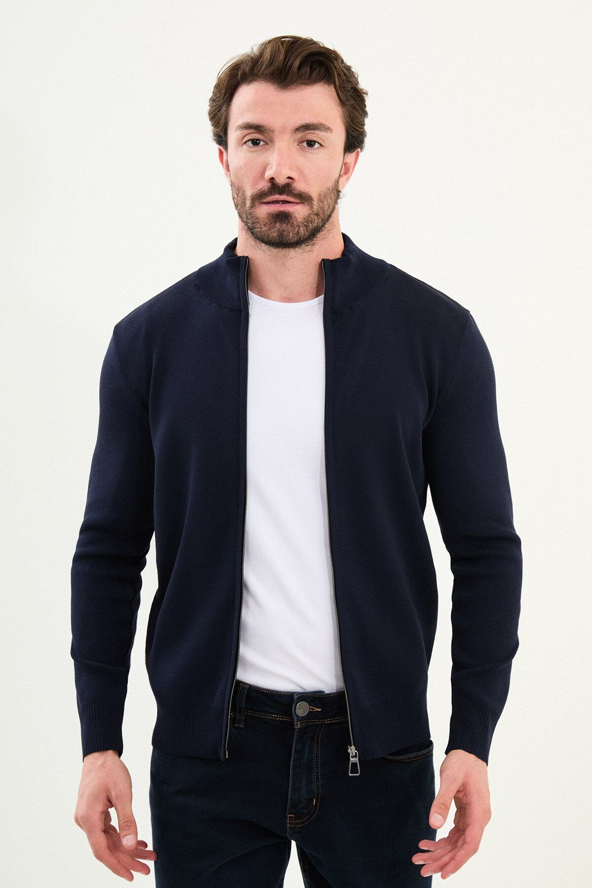 A man wearing a slim fit navy blue zip cardigan with a stand-up collar, styled with a white T-shirt and dark jeans.