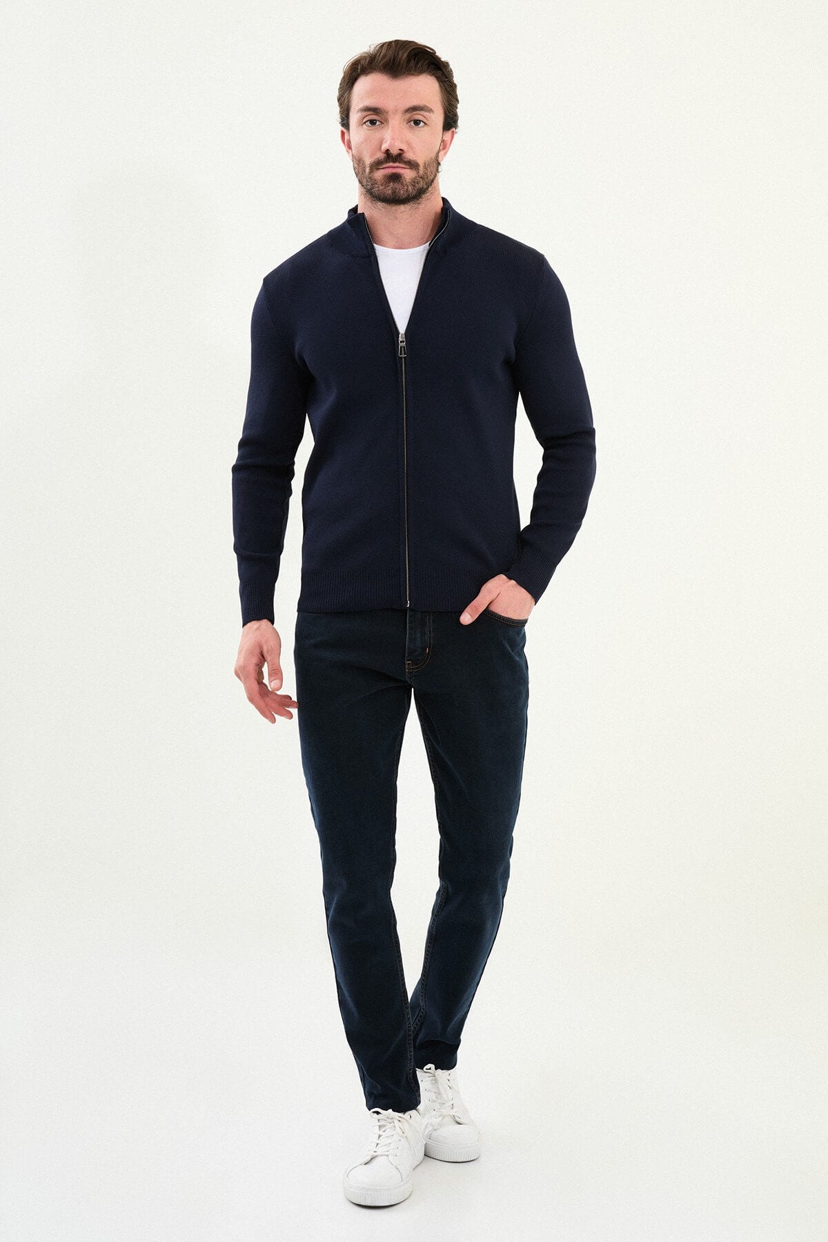 A man wearing a slim fit navy blue zip cardigan with a stand-up collar, styled with a white T-shirt and dark jeans.