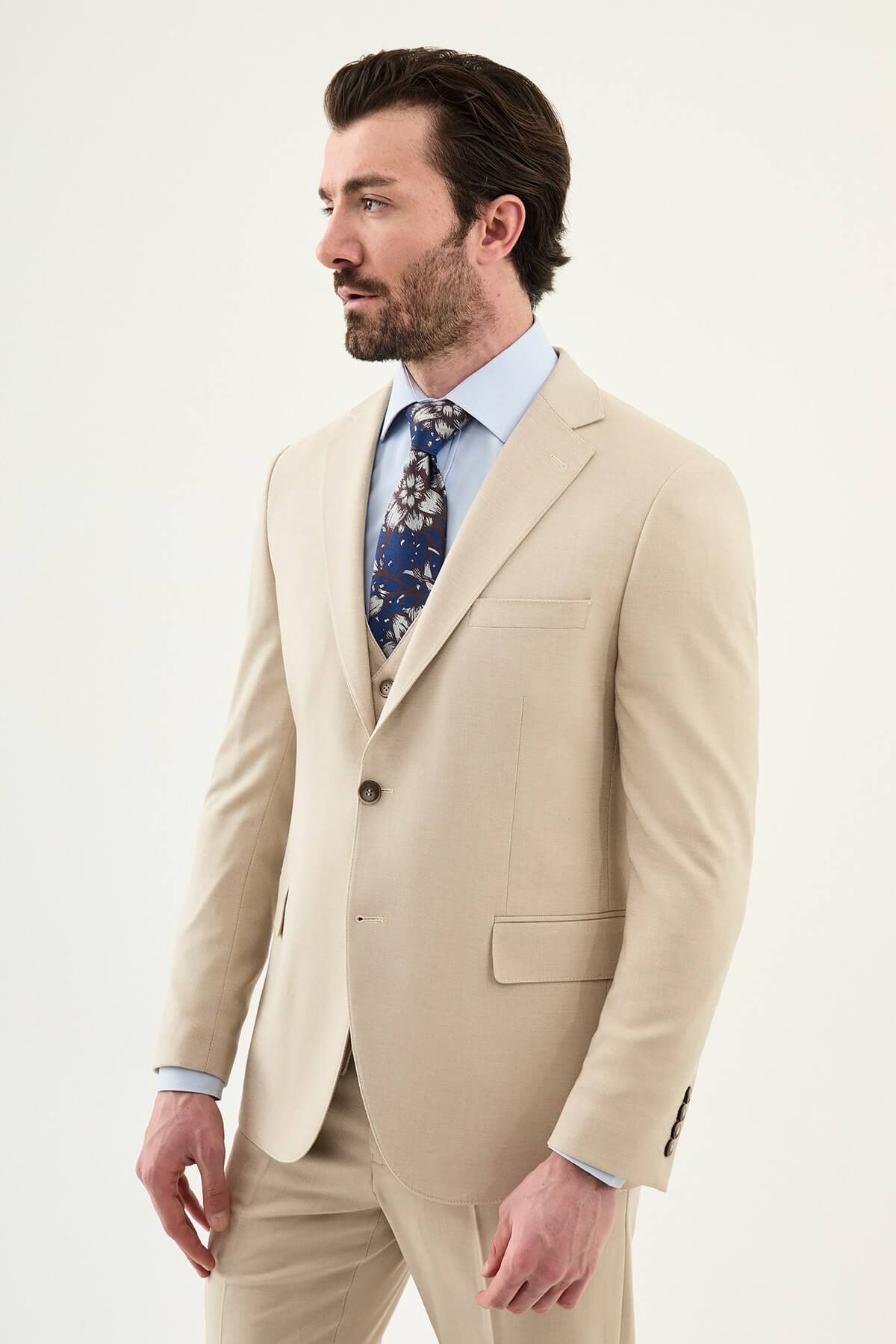 Model wearing a beige slim-fit cotton-linen three-piece suit with a pale blue dress shirt, navy floral tie, and matching tailored trousers.