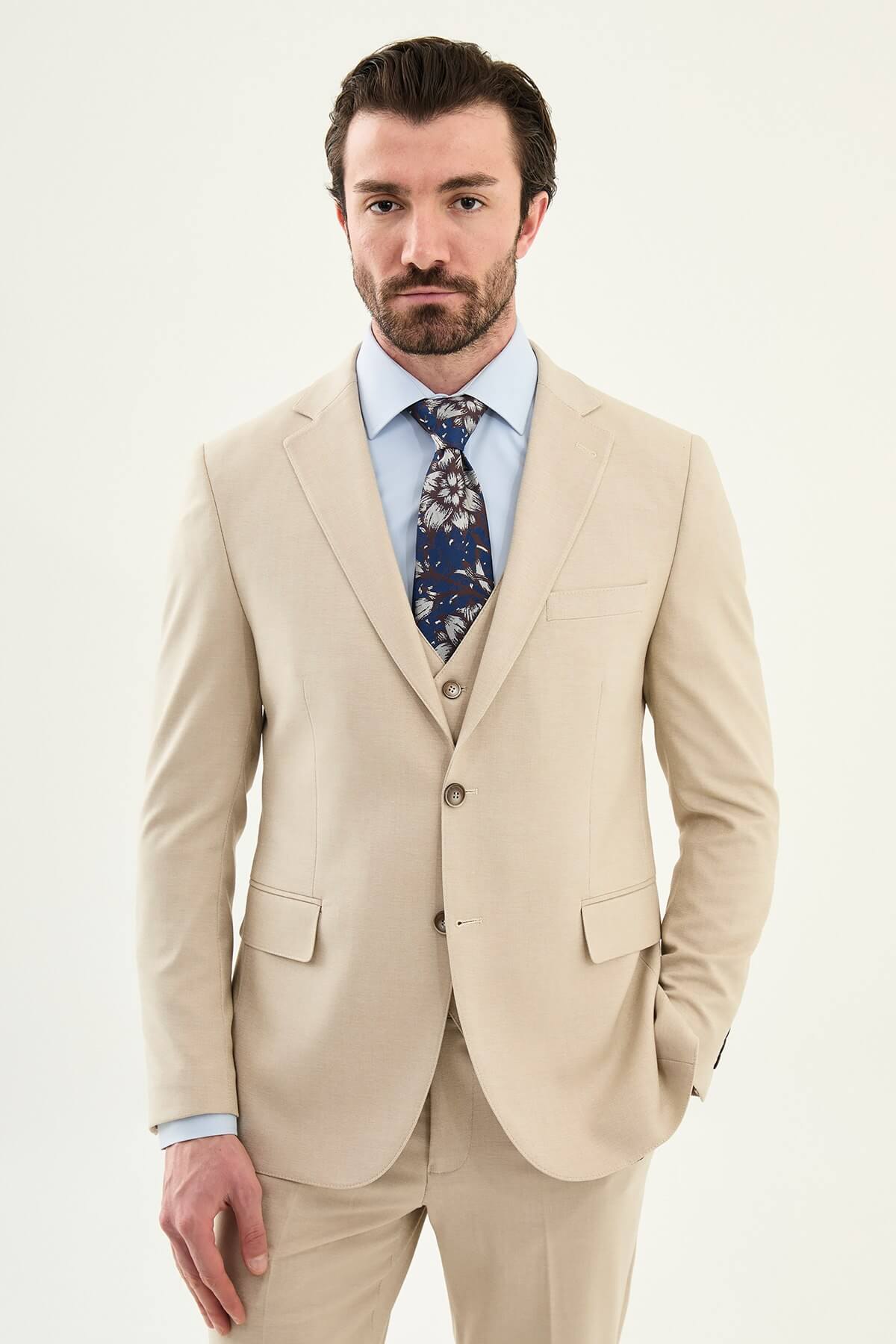 Model wearing a beige slim-fit cotton-linen three-piece suit with a pale blue dress shirt, navy floral tie, and matching tailored trousers.