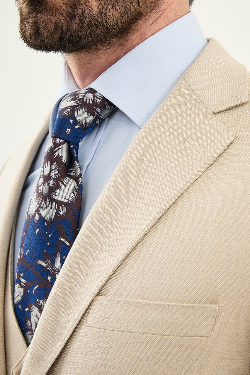 Model wearing a beige slim-fit cotton-linen three-piece suit with a pale blue dress shirt, navy floral tie, and matching tailored trousers.