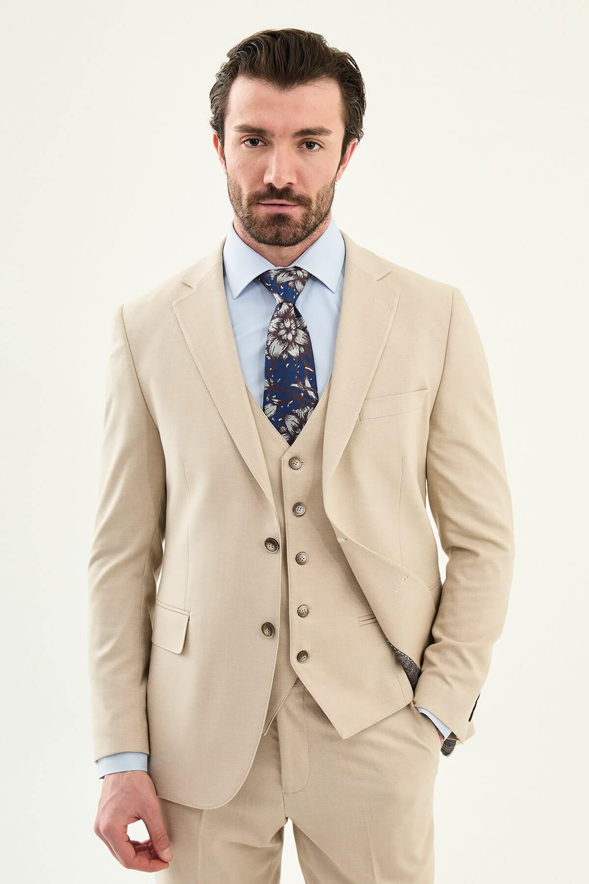 Model wearing a beige slim-fit cotton-linen three-piece suit with a pale blue dress shirt, navy floral tie, and matching tailored trousers.