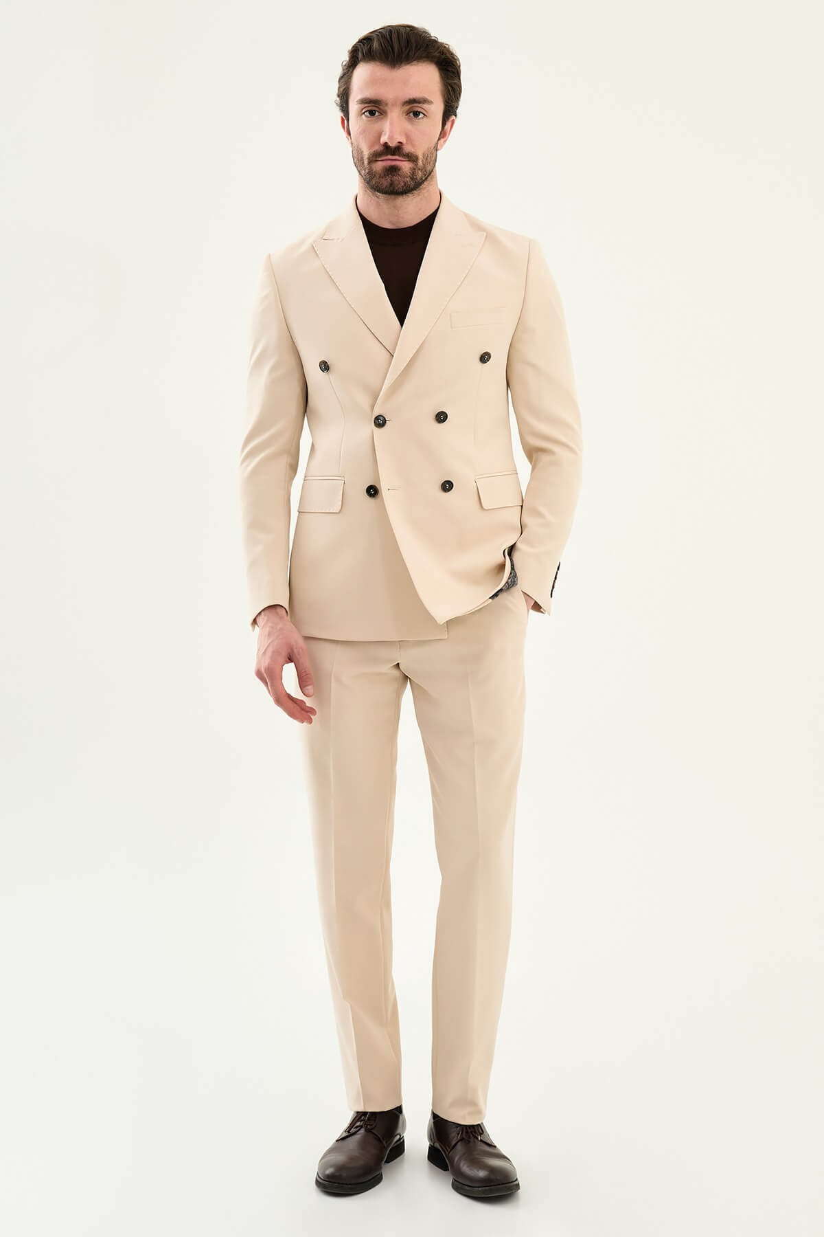 Model wearing a beige slim-fit double-breasted suit with peak lapels, dark buttons, matching trousers, a dark brown knit top, and brown leather dress shoes.