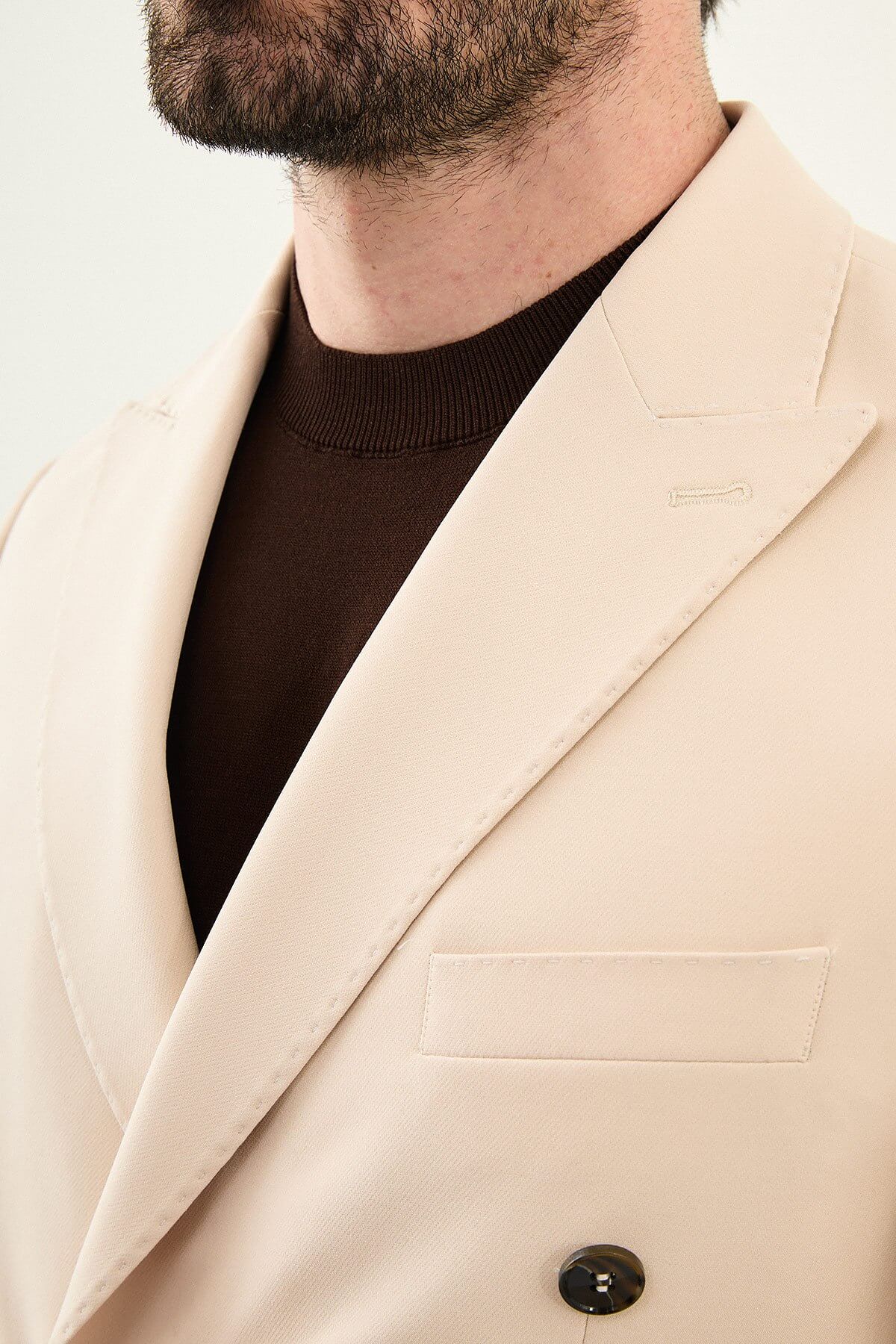 Model wearing a beige slim-fit double-breasted suit with peak lapels, dark buttons, matching trousers, a dark brown knit top, and brown leather dress shoes.