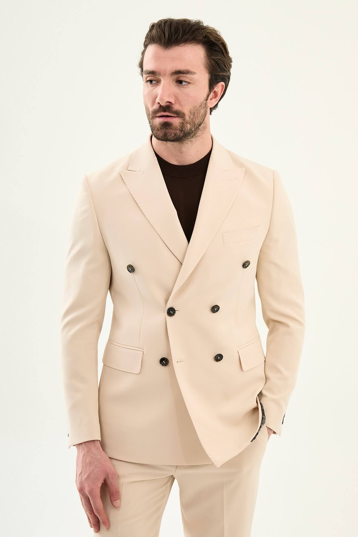 Model wearing a beige slim-fit double-breasted suit with peak lapels, dark buttons, matching trousers, a dark brown knit top, and brown leather dress shoes.