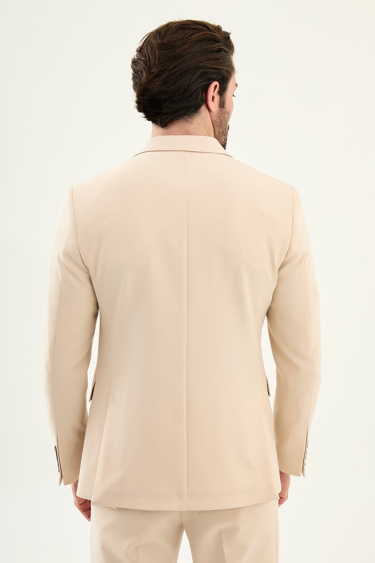 Model wearing a beige slim-fit double-breasted suit with peak lapels, dark buttons, matching trousers, a dark brown knit top, and brown leather dress shoes.