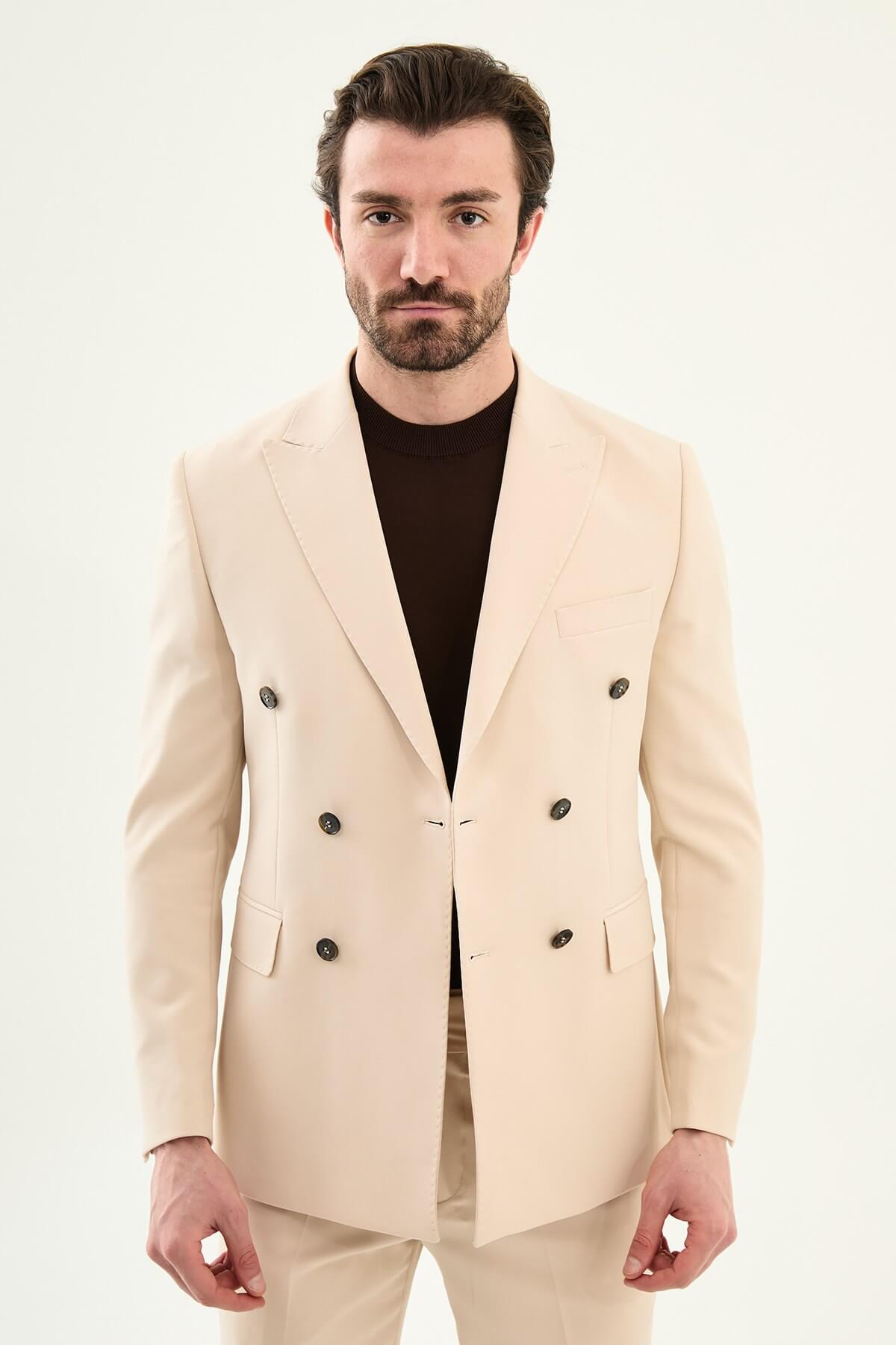 Model wearing a beige slim-fit double-breasted suit with peak lapels, dark buttons, matching trousers, a dark brown knit top, and brown leather dress shoes.