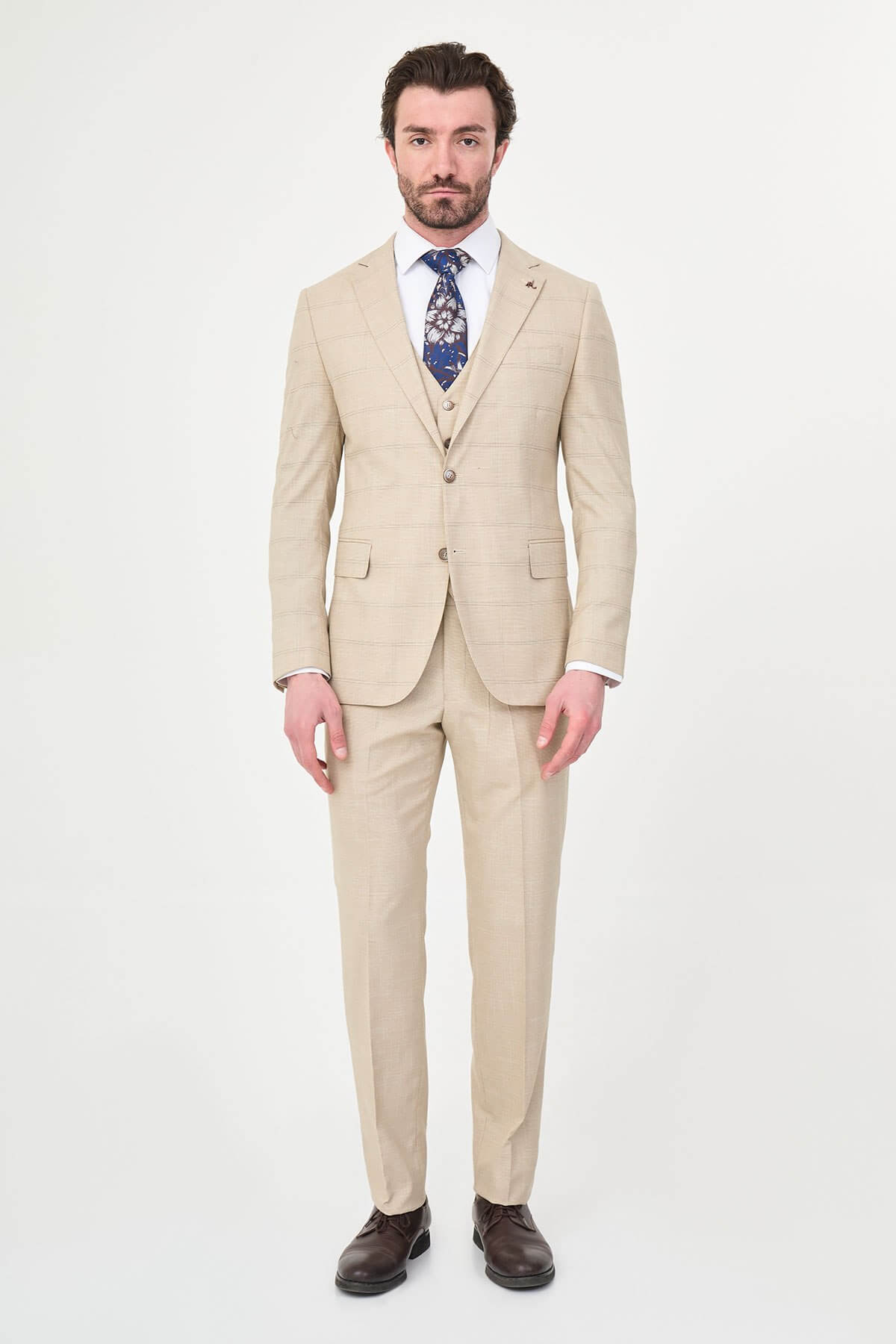 a beige check slim-fit three-piece suit with matching waistcoat and trousers, white dress shirt, patterned navy and brown tie, and dark brown leather shoes.