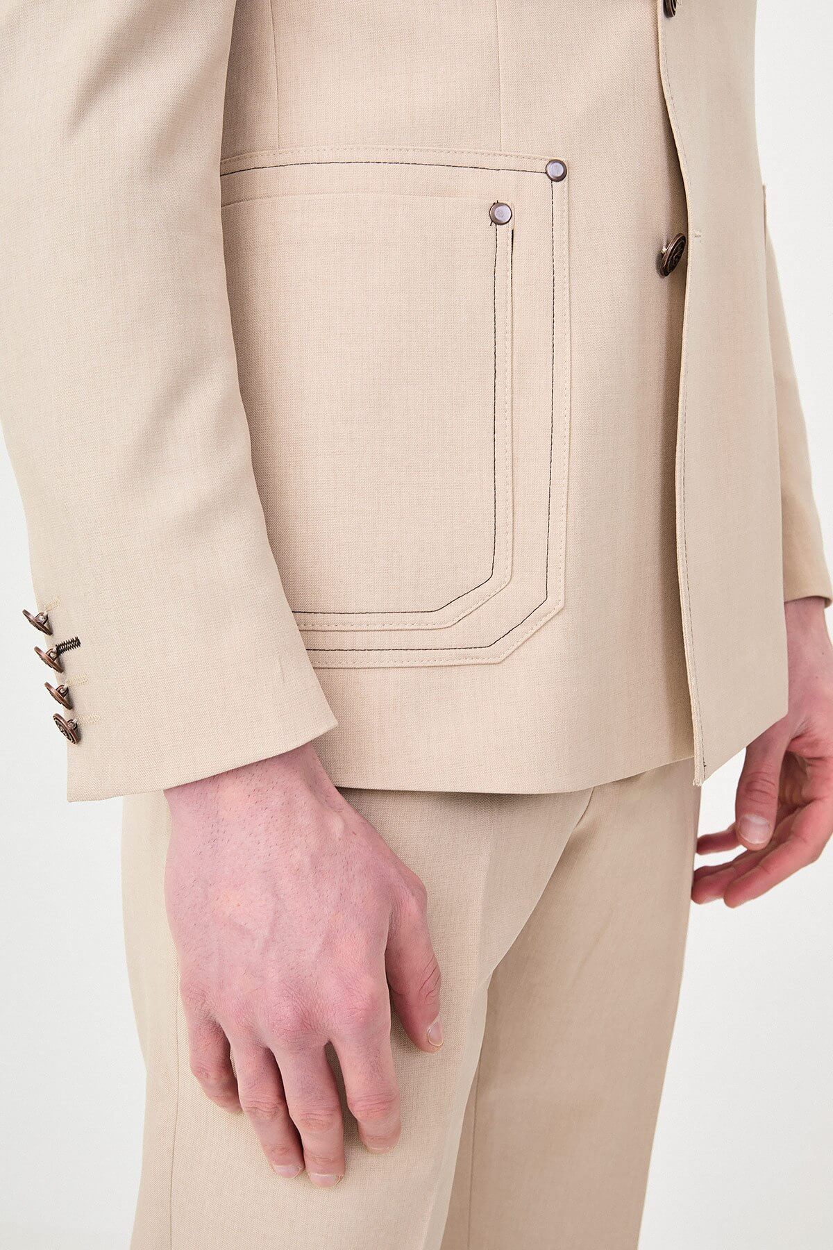 Model wearing a beige slim-fit two-piece wedding suit with notch lapels, tonal stitched patch pockets, matching trousers, olive knitwear, and dark brown leather shoes.