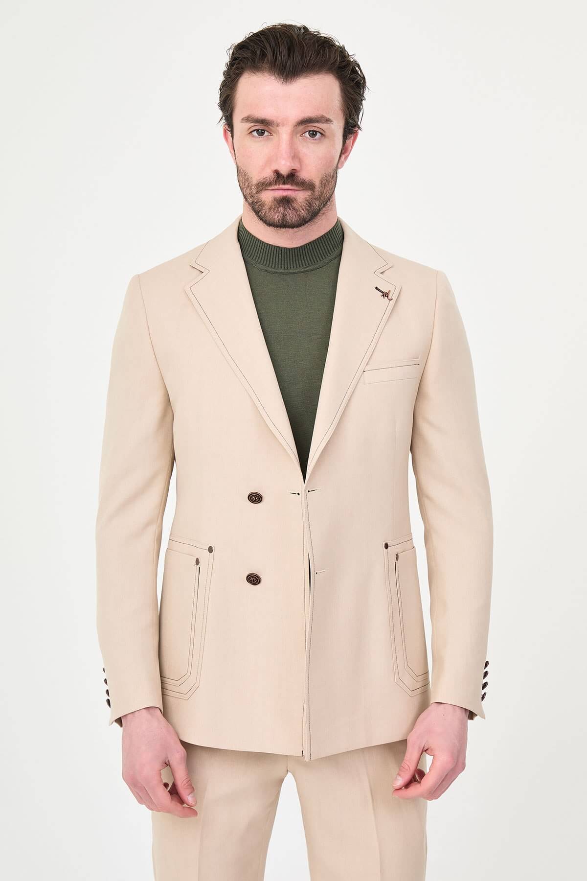 Model wearing a beige slim-fit two-piece wedding suit with notch lapels, tonal stitched patch pockets, matching trousers, olive knitwear, and dark brown leather shoes.