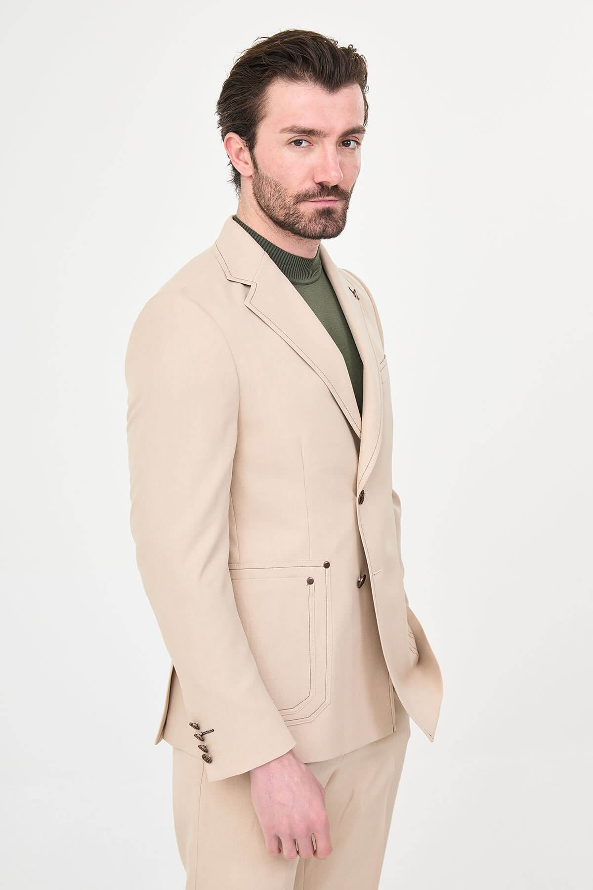 Model wearing a beige slim-fit two-piece wedding suit with notch lapels, tonal stitched patch pockets, matching trousers, olive knitwear, and dark brown leather shoes.
