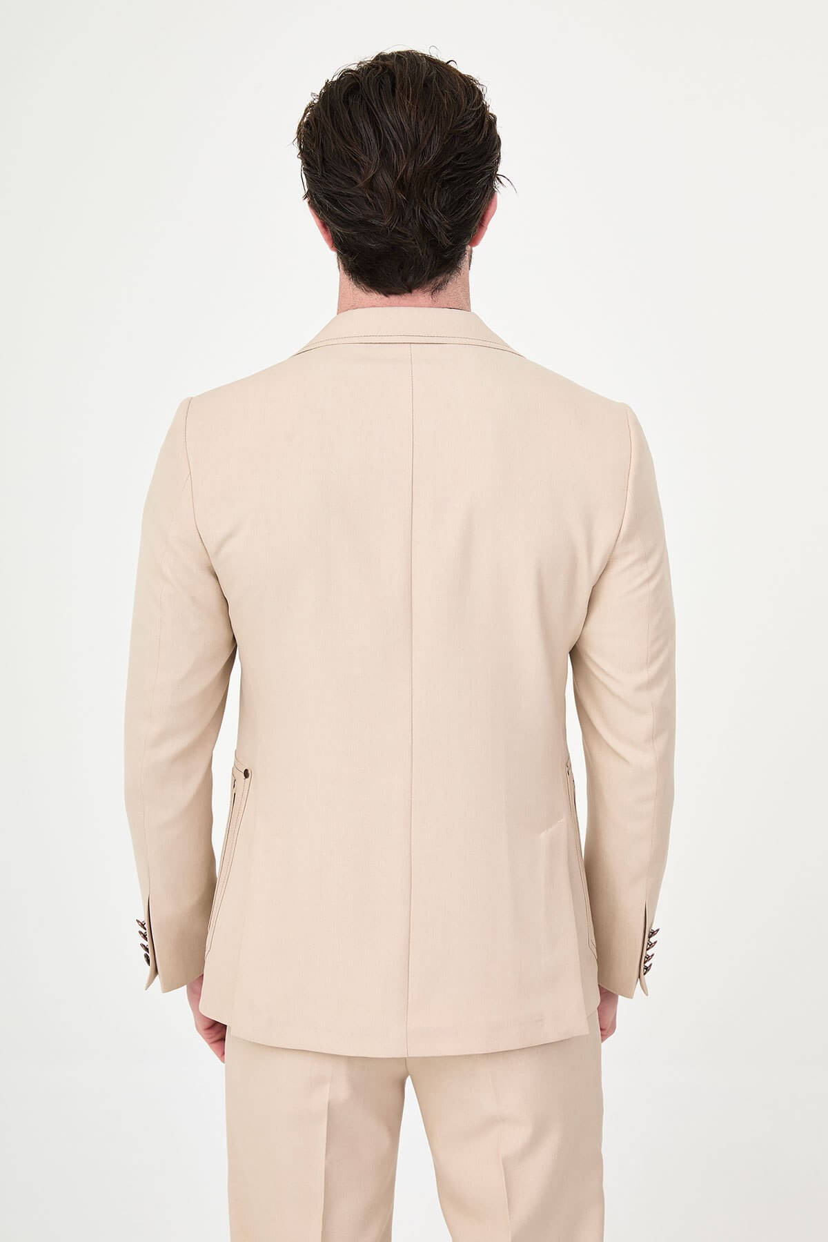 Model wearing a beige slim-fit two-piece wedding suit with notch lapels, tonal stitched patch pockets, matching trousers, olive knitwear, and dark brown leather shoes.