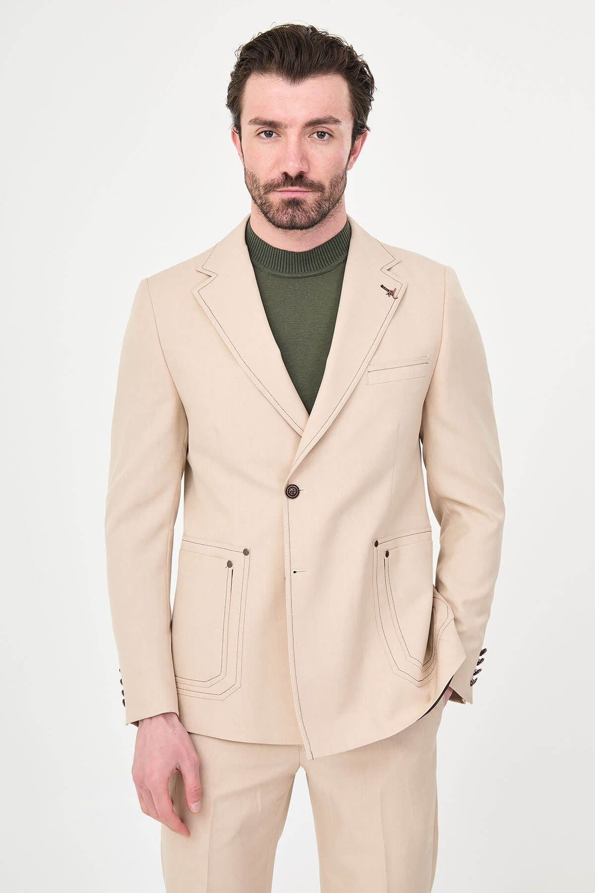Model wearing a beige slim-fit two-piece wedding suit with notch lapels, tonal stitched patch pockets, matching trousers, olive knitwear, and dark brown leather shoes.