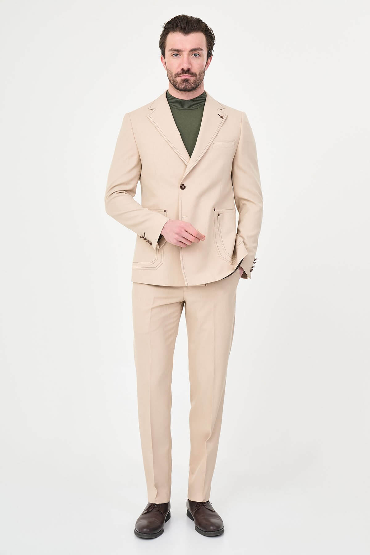 Model wearing a beige slim-fit two-piece wedding suit with notch lapels, tonal stitched patch pockets, matching trousers, olive knitwear, and dark brown leather shoes.