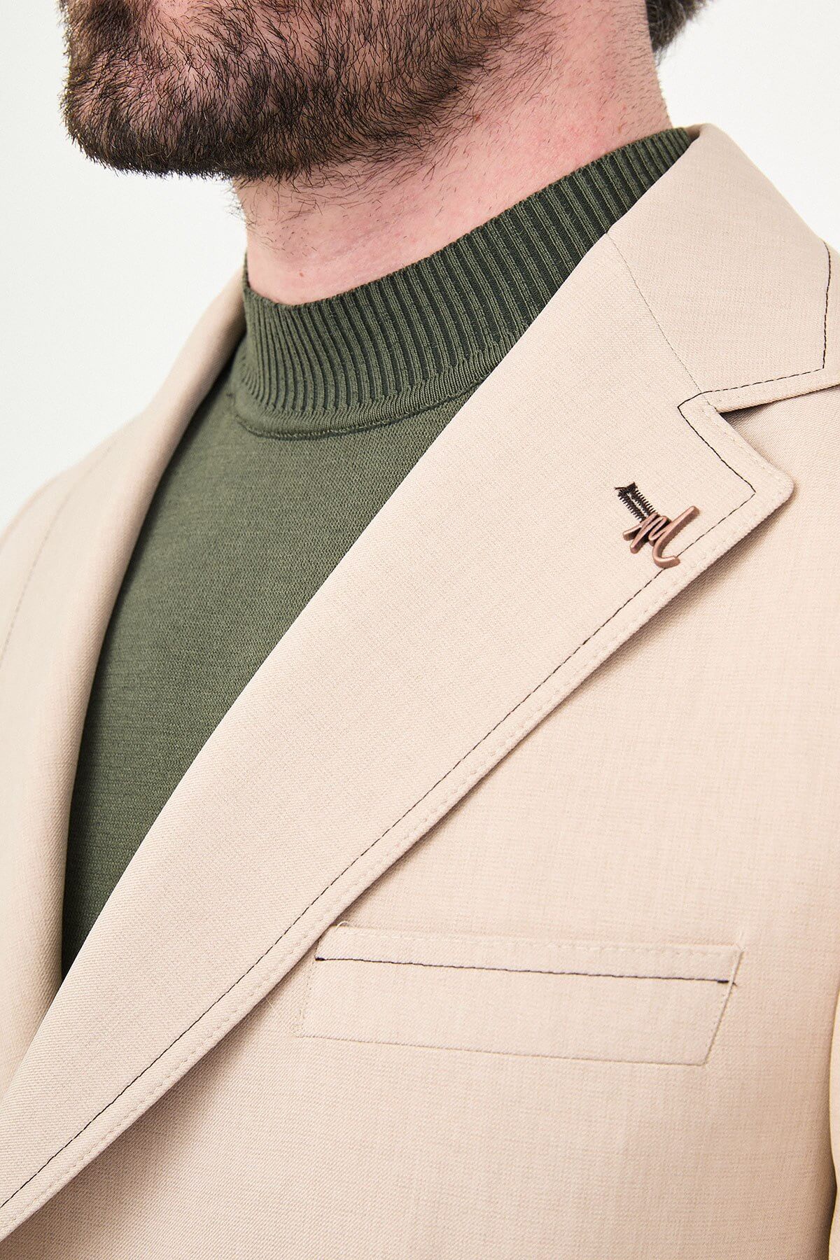 Model wearing a beige slim-fit two-piece wedding suit with notch lapels, tonal stitched patch pockets, matching trousers, olive knitwear, and dark brown leather shoes.