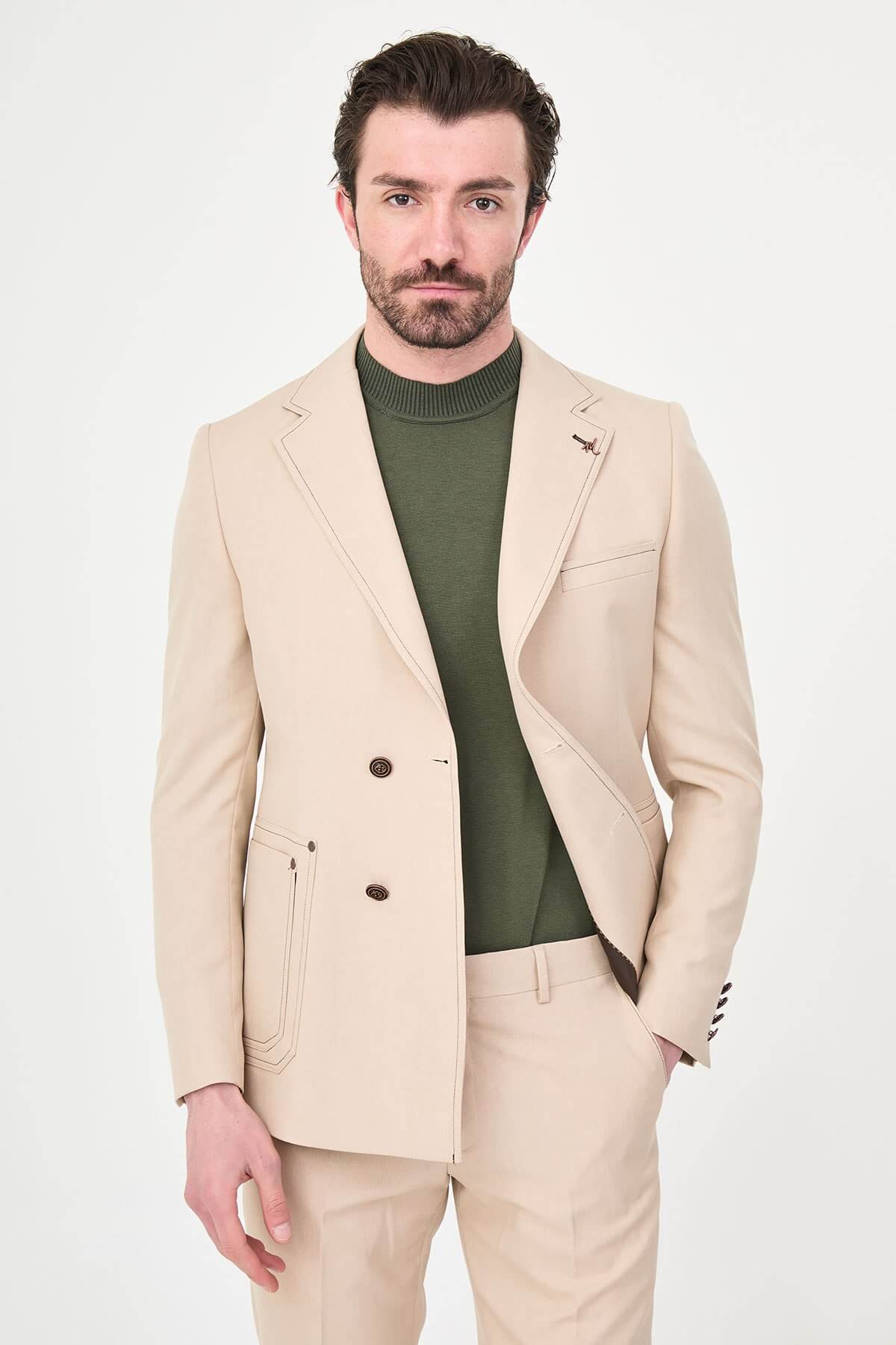 Model wearing a beige slim-fit two-piece wedding suit with notch lapels, tonal stitched patch pockets, matching trousers, olive knitwear, and dark brown leather shoes.