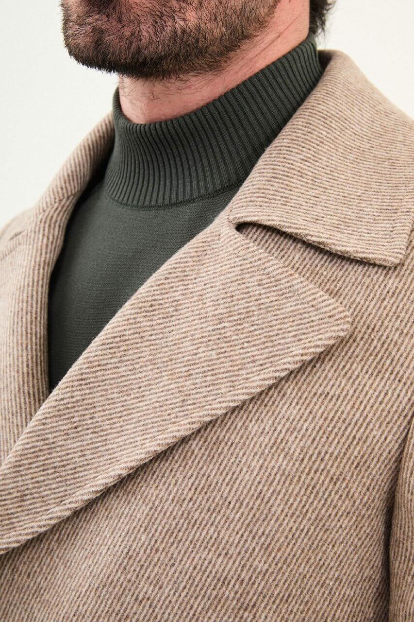 A man wearing a beige double-breasted slim fit peacoat with wide lapels, styled over a dark green turtleneck and paired with dark trousers