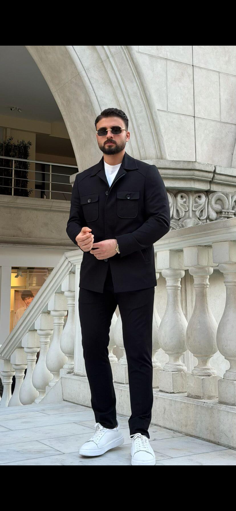 Men’s black safari co-set with buttoned jacket and multiple pockets, paired with black pants and white t-shirt, worn casually outdoors.