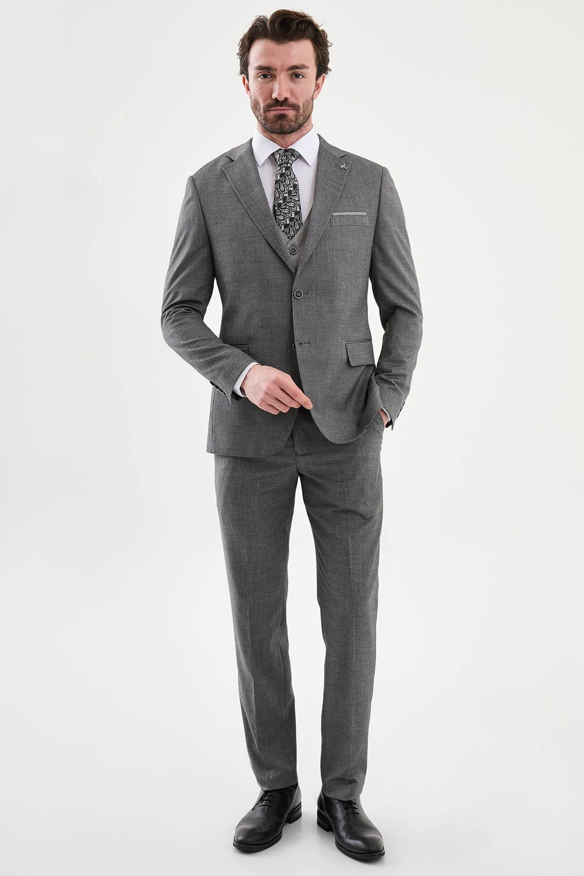 Model wearing a charcoal gray slim-fit three-piece suit with a white dress shirt, patterned black tie, matching waistcoat, tailored trousers, and black leather shoes.