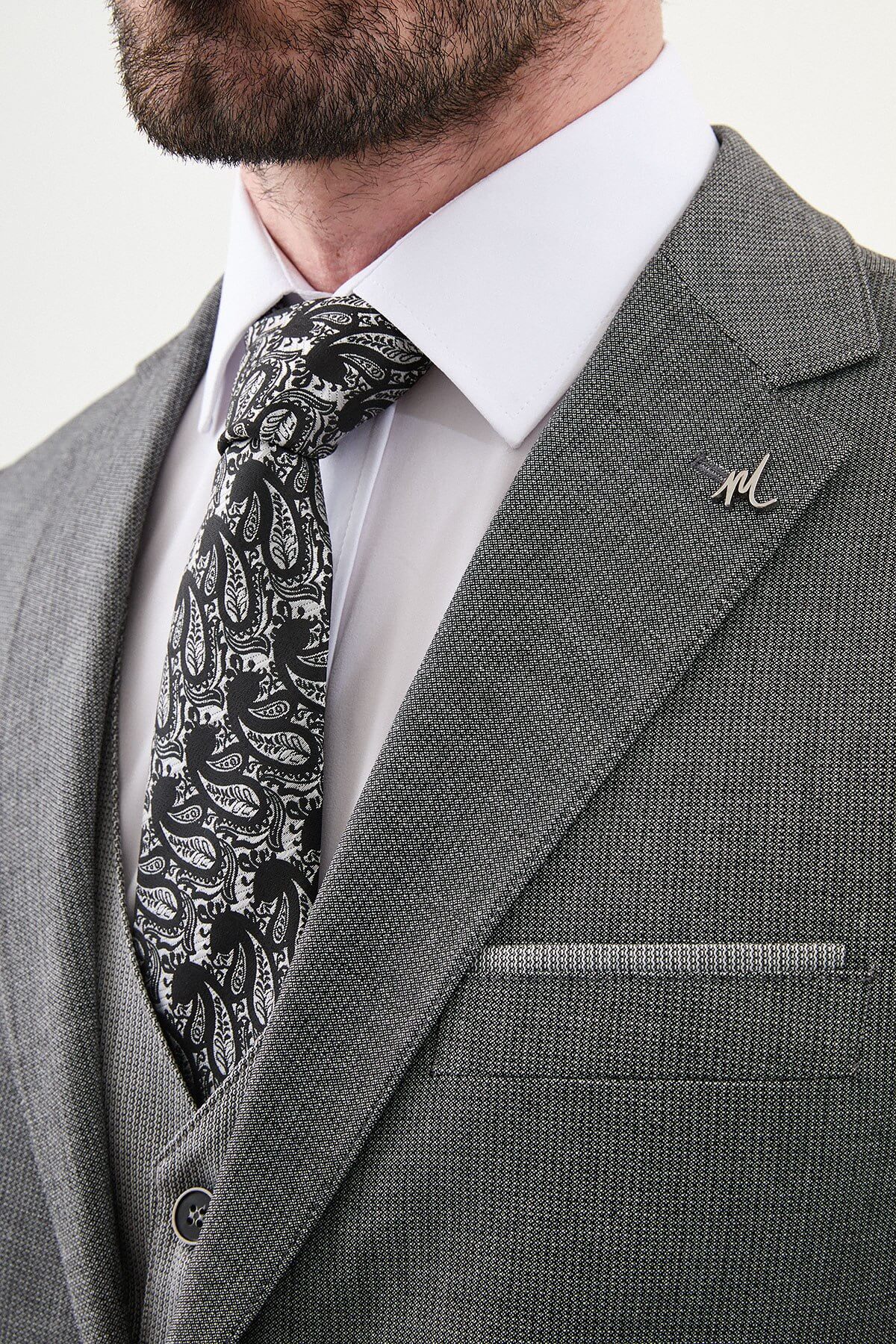 Model wearing a charcoal gray slim-fit three-piece suit with a white dress shirt, patterned black tie, matching waistcoat, tailored trousers, and black leather shoes.