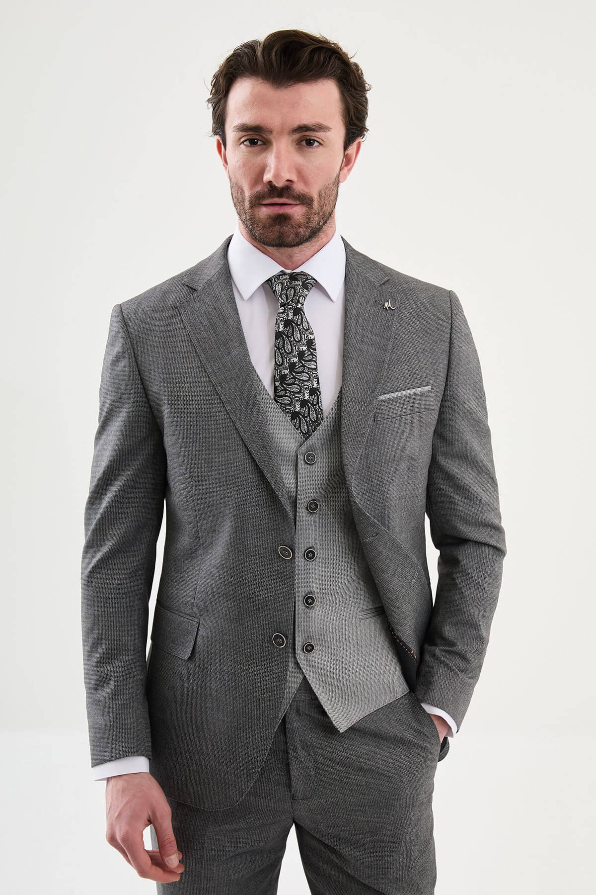 Model wearing a charcoal gray slim-fit three-piece suit with a white dress shirt, patterned black tie, matching waistcoat, tailored trousers, and black leather shoes.