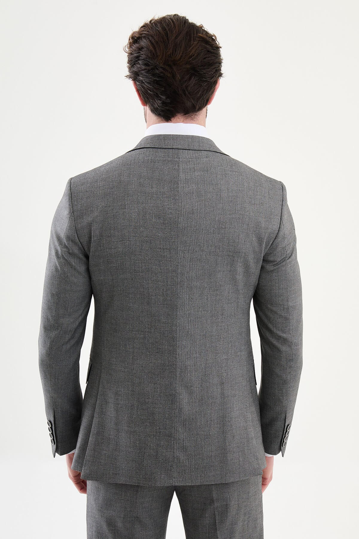 Model wearing a charcoal gray slim-fit three-piece suit with a white dress shirt, patterned black tie, matching waistcoat, tailored trousers, and black leather shoes.
