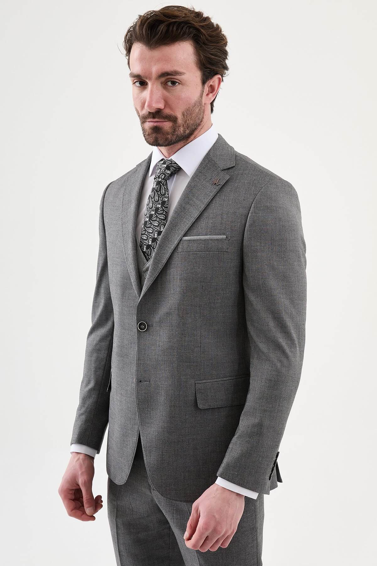 Model wearing a charcoal gray slim-fit three-piece suit with a white dress shirt, patterned black tie, matching waistcoat, tailored trousers, and black leather shoes.