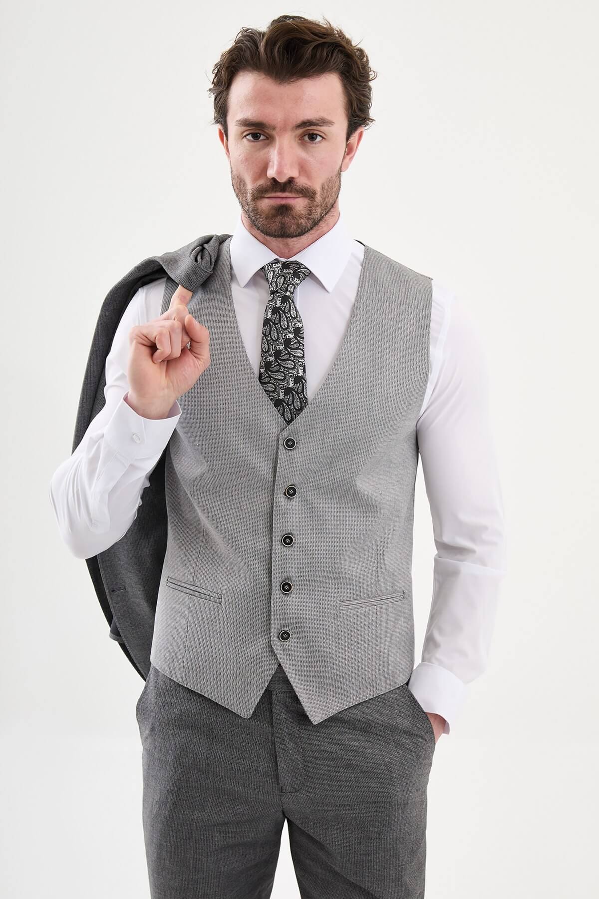Model wearing a charcoal gray slim-fit three-piece suit with a white dress shirt, patterned black tie, matching waistcoat, tailored trousers, and black leather shoes.