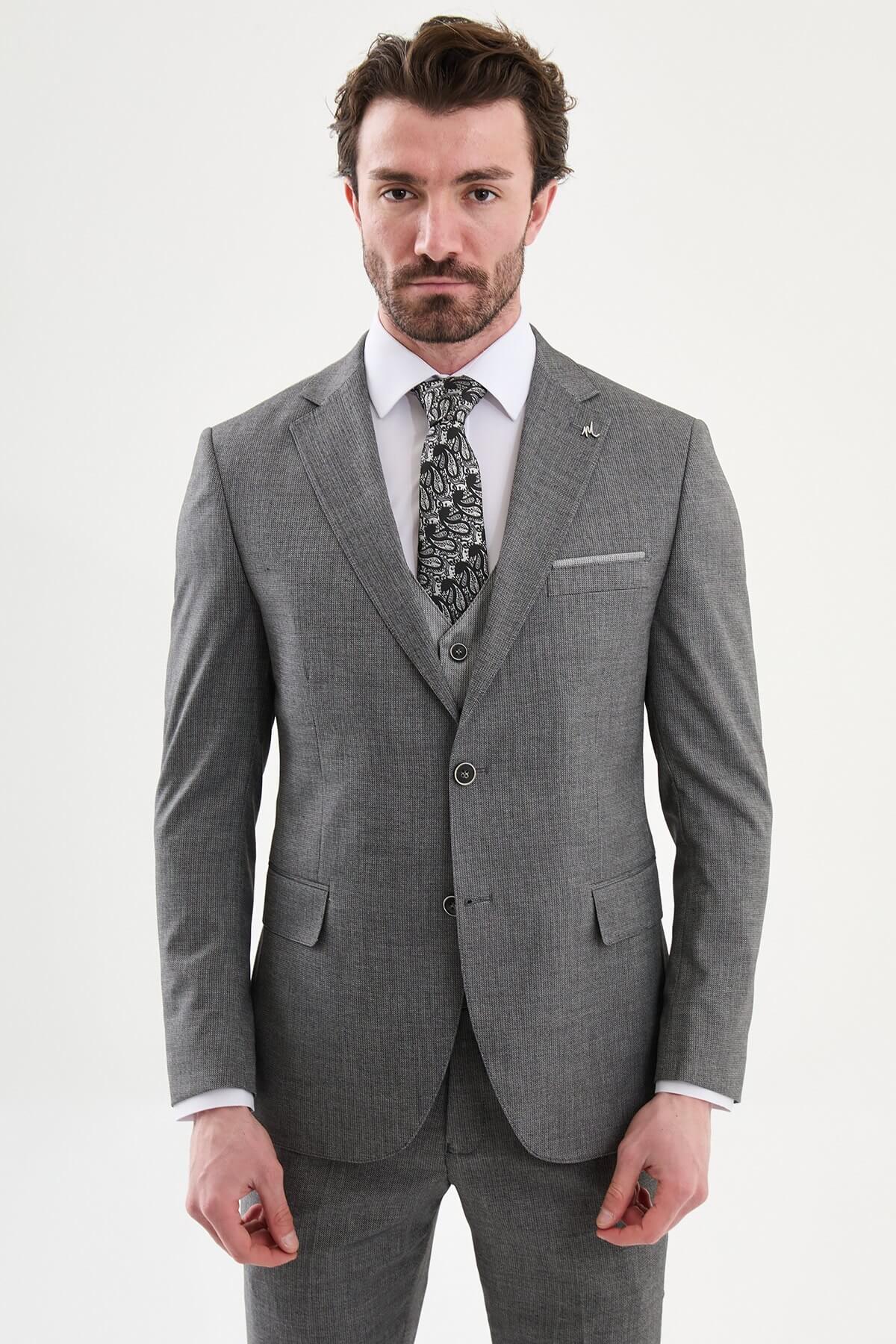 Model wearing a charcoal gray slim-fit three-piece suit with a white dress shirt, patterned black tie, matching waistcoat, tailored trousers, and black leather shoes.