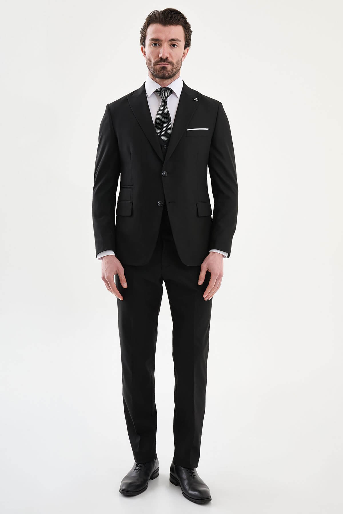man wearing a black slim-fit three-piece suit, including blazer, waistcoat, and trousers, styled with a white dress shirt and black tie, suitable for formal events or weddings.