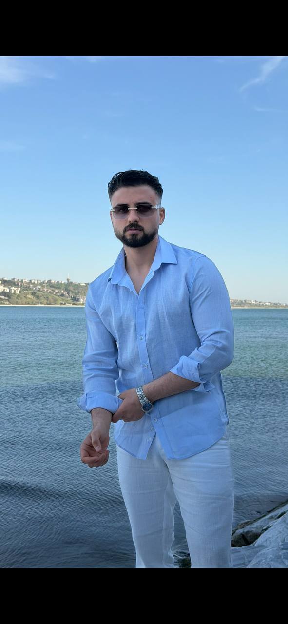 man wearing a light blue linen collared shirt, standing on a beach with rolled-up sleeves, paired with white trousers.