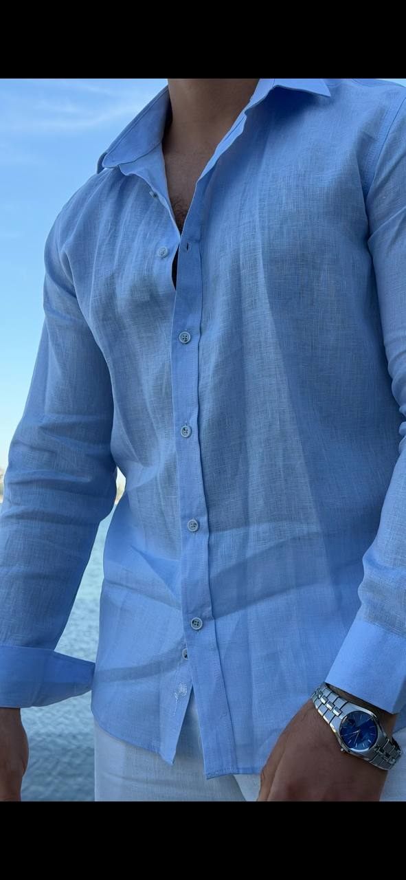 man wearing a light blue linen collared shirt, standing on a beach with rolled-up sleeves, paired with white trousers.