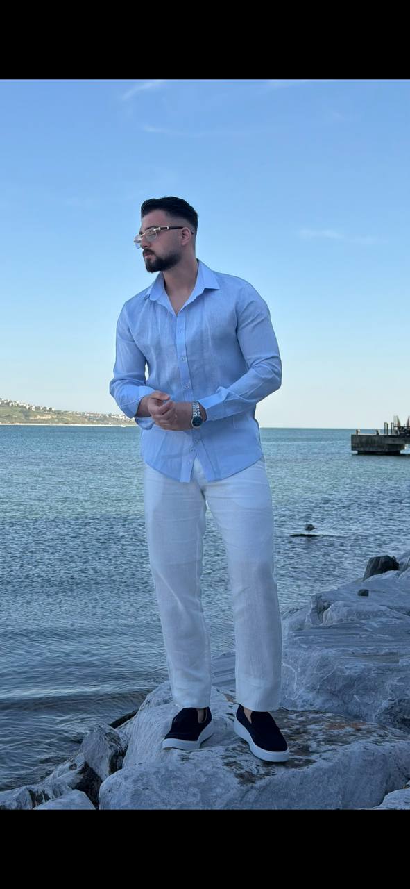 man wearing a light blue linen collared shirt, standing on a beach with rolled-up sleeves, paired with white trousers.
