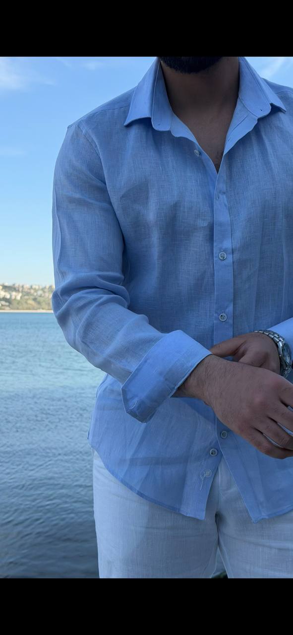 man wearing a light blue linen collared shirt, standing on a beach with rolled-up sleeves, paired with white trousers.