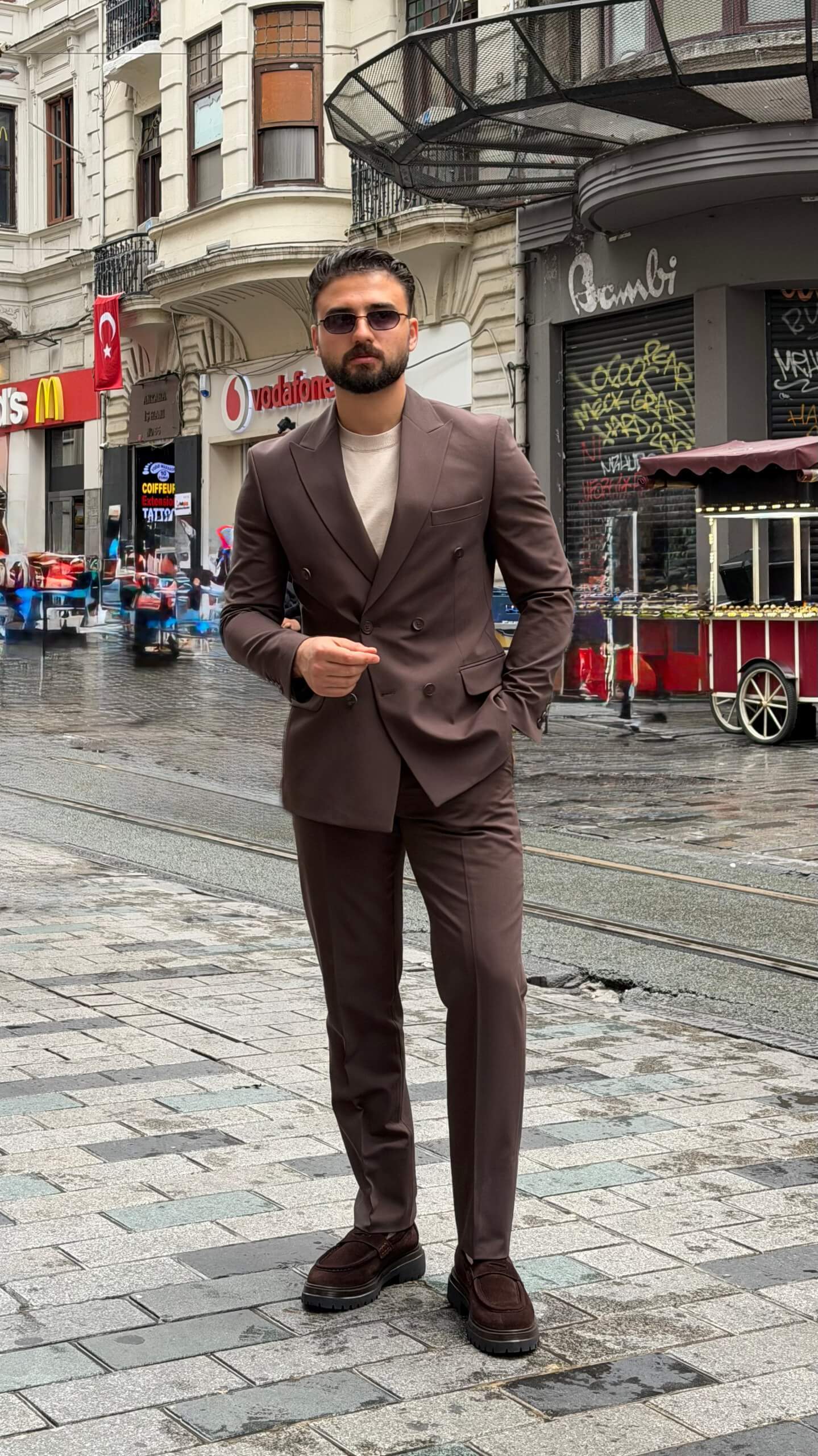 Man wearing a brown double-breasted slim-fit suit with matching trousers, styled with a beige shirt and patterned tie, standing against a neutral background.