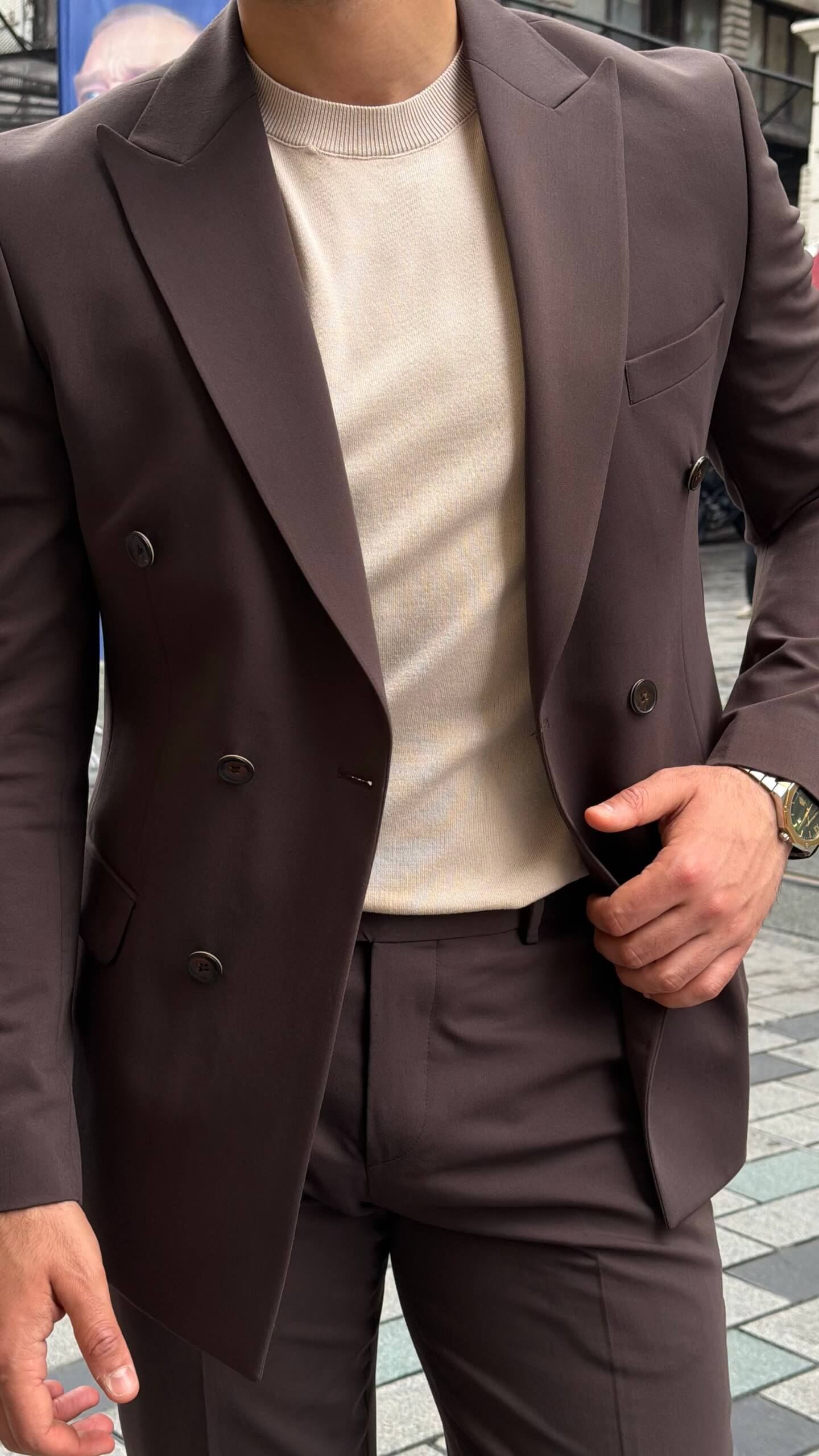 Man wearing a brown double-breasted slim-fit suit with matching trousers, styled with a beige shirt and patterned tie, standing against a neutral background.