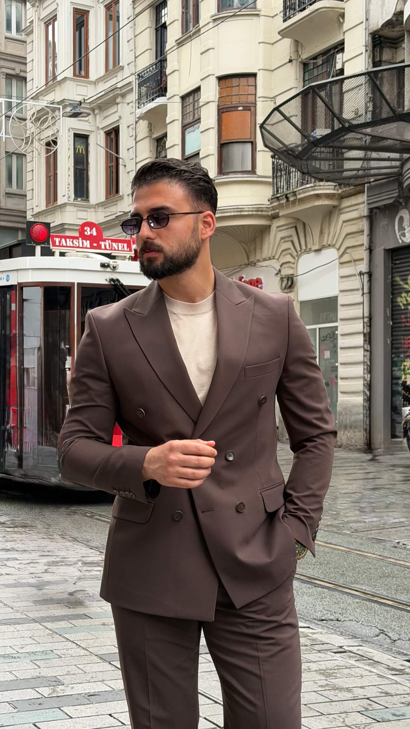 Man wearing a brown double-breasted slim-fit suit with matching trousers, styled with a beige shirt and patterned tie, standing against a neutral background.