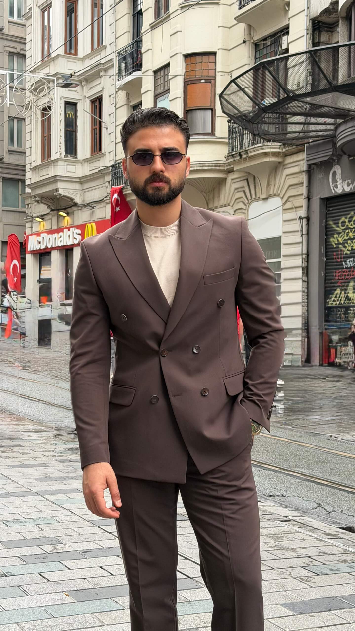 Man wearing a brown double-breasted slim-fit suit with matching trousers, styled with a beige shirt and patterned tie, standing against a neutral background.