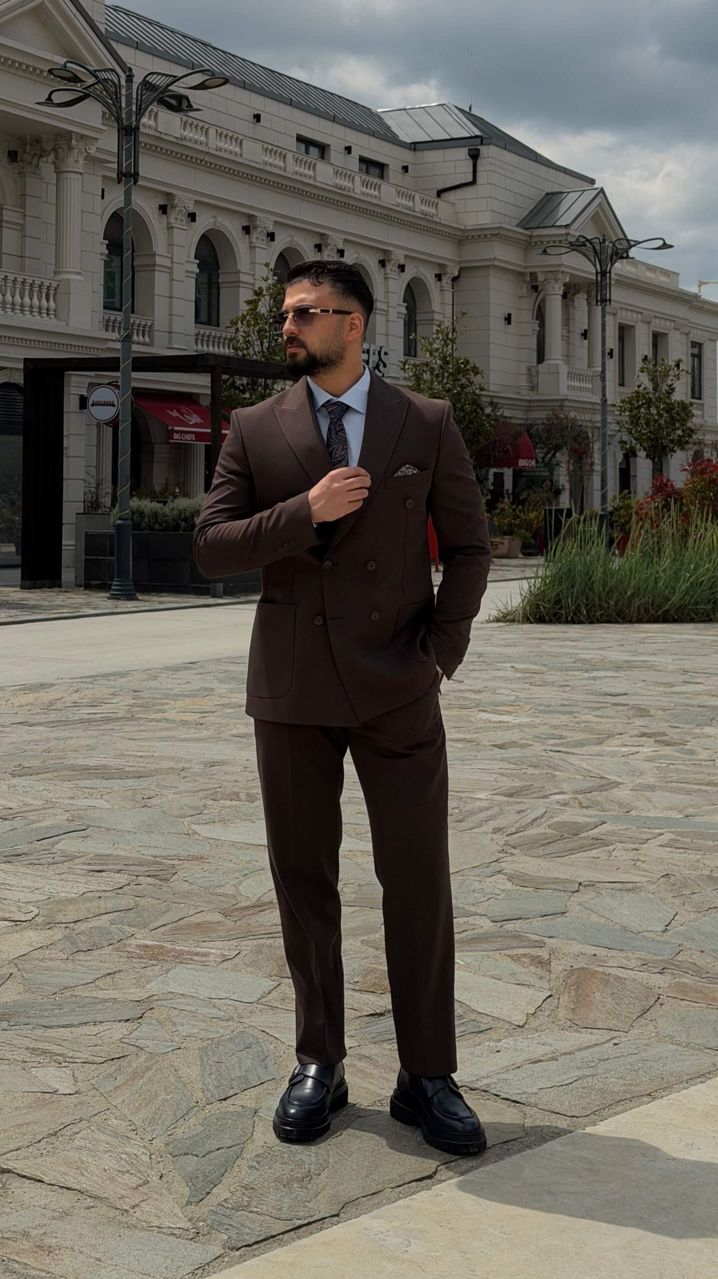 Men wearing a brown double-breasted slim-fit two-piece suit standing outdoors, styled with black shoes and a tie.