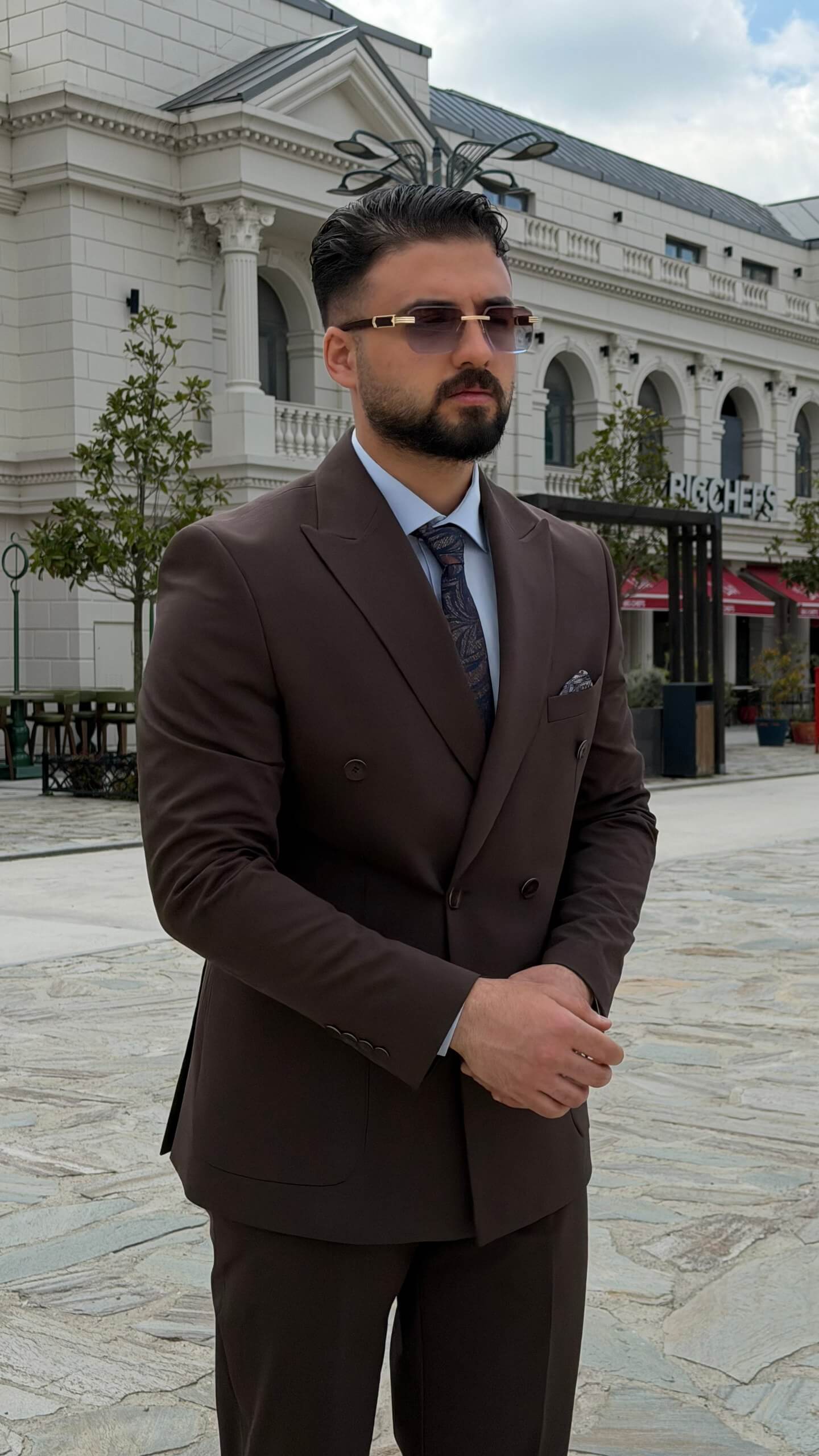 Men wearing a brown double-breasted slim-fit two-piece suit standing outdoors, styled with black shoes and a tie.