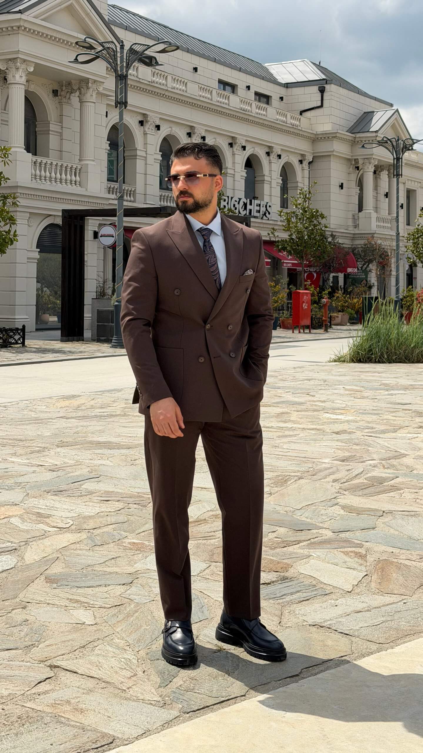 Men wearing a brown double-breasted slim-fit two-piece suit standing outdoors, styled with black shoes and a tie.