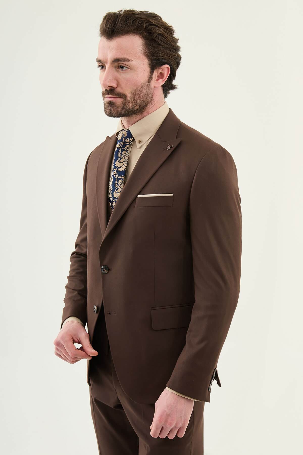 Man wearing a brown three-piece suit with blazer, waistcoat, and trousers, paired with a beige shirt and patterned tie