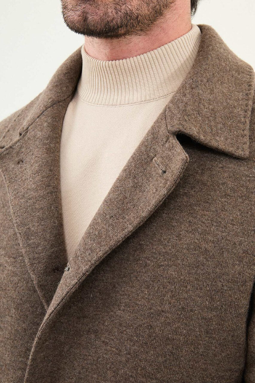 A man wearing a slim fit brown coat over a beige turtleneck, styled with brown trousers and dark brown Chelsea boots.