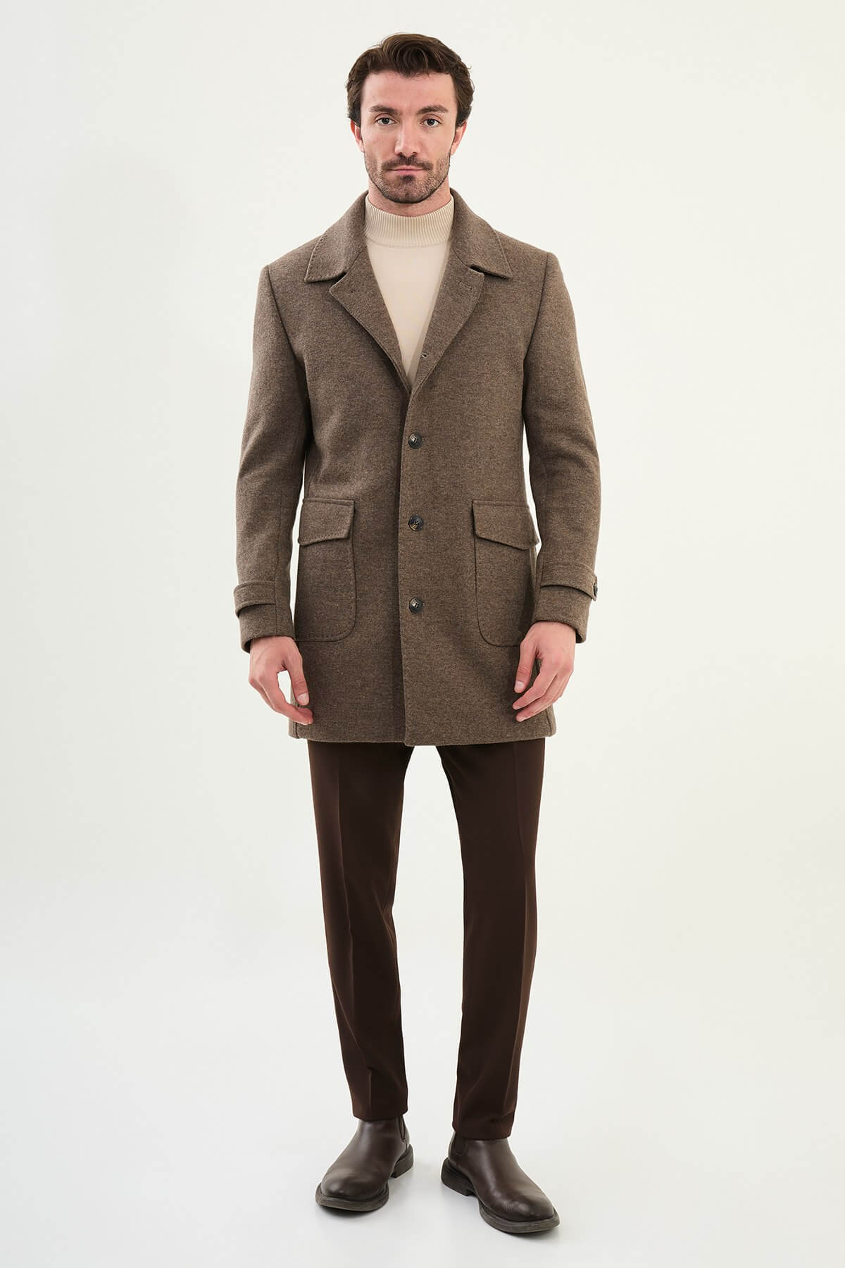 A man wearing a slim fit brown coat over a beige turtleneck, styled with brown trousers and dark brown Chelsea boots.