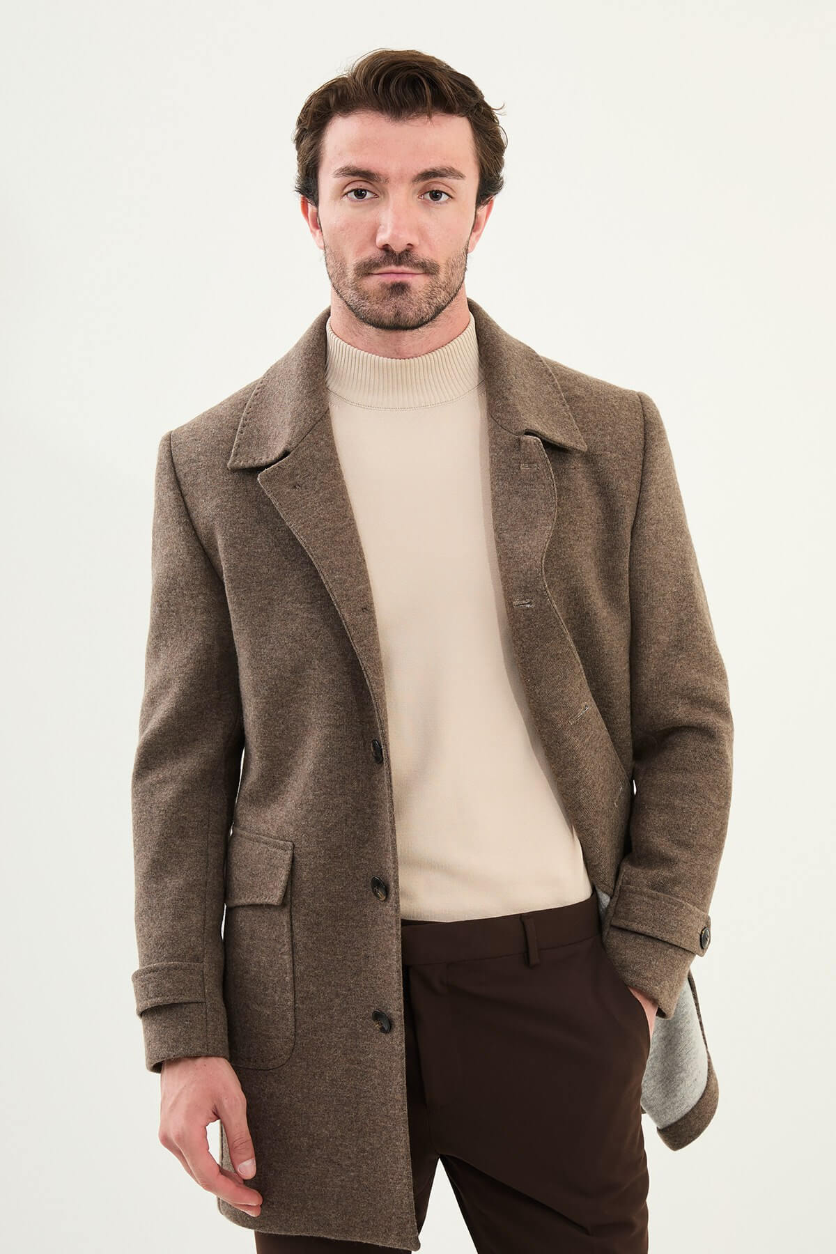 A man wearing a slim fit brown coat over a beige turtleneck, styled with brown trousers and dark brown Chelsea boots.