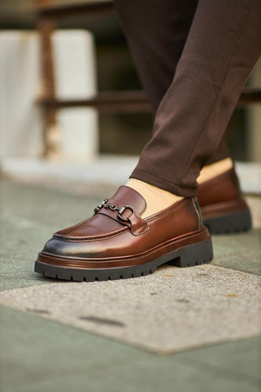 Brown leather bit loafers with chunky sole worn with brown trousers on outdoor pavement.
