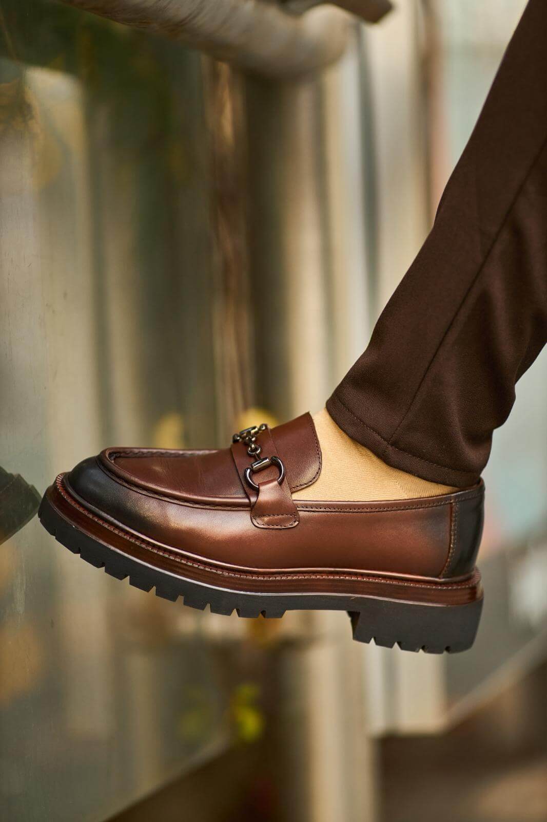 Brown leather bit loafers with chunky sole worn with brown trousers on outdoor pavement.
